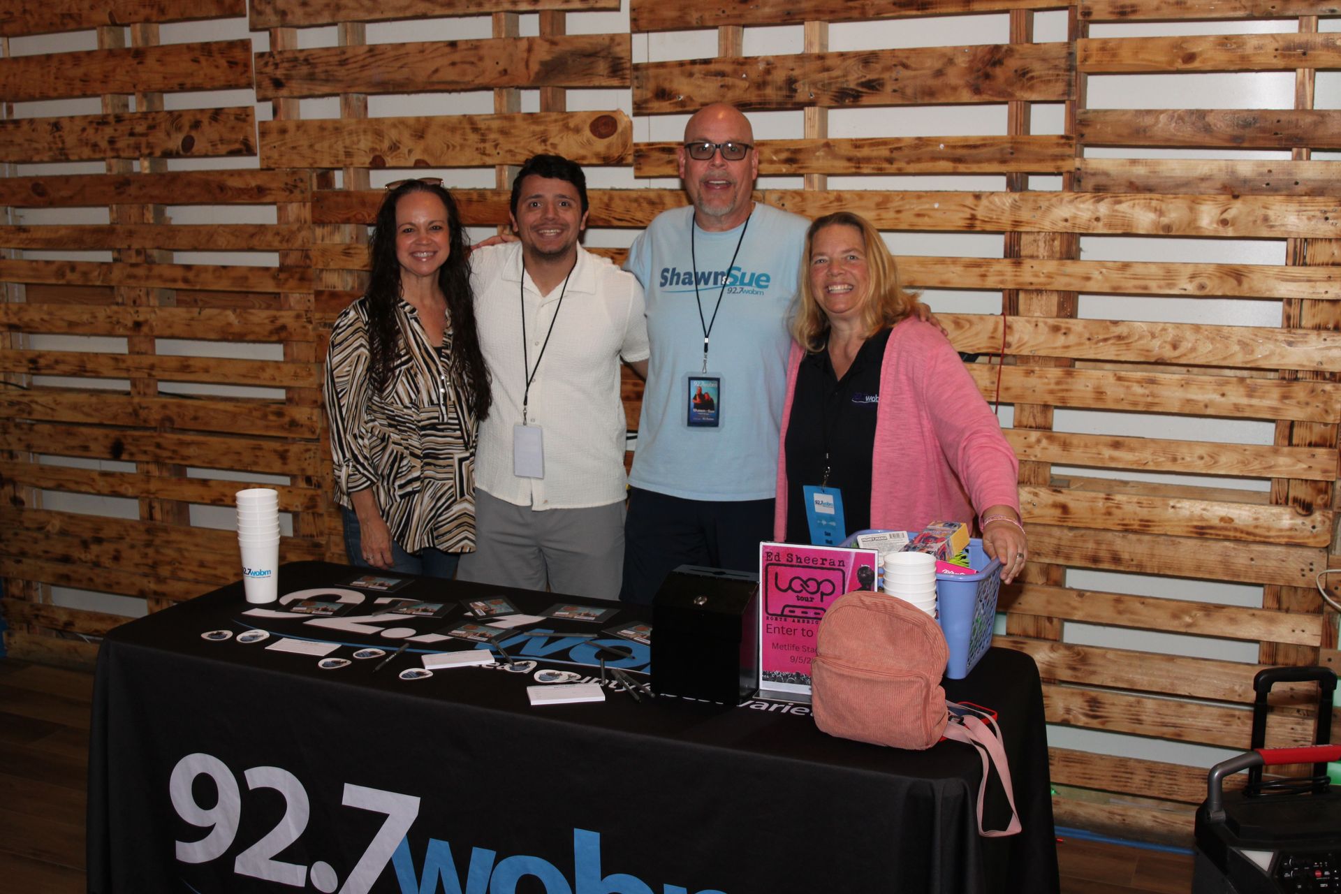 Four people behind a 92.7 WOBM booth with a pink sign and table display at an indoor event.