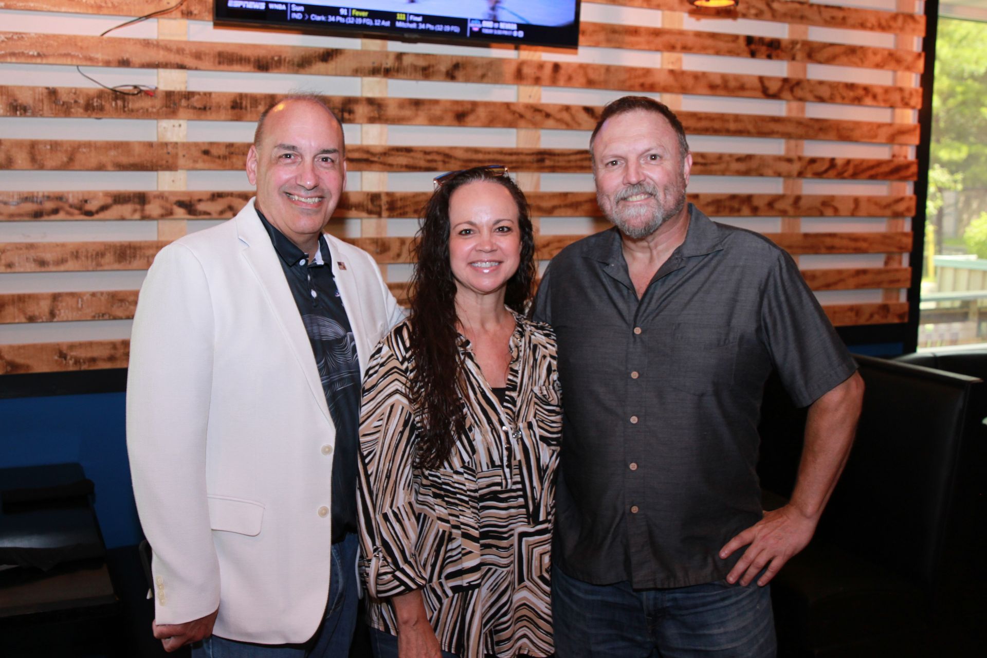 Three people standing together indoors, smiling in front of a wood slat wall.