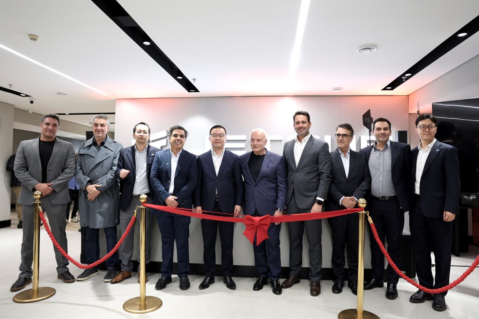 Group of people in suits posing behind a red ribbon at an indoor ribbon-cutting event.