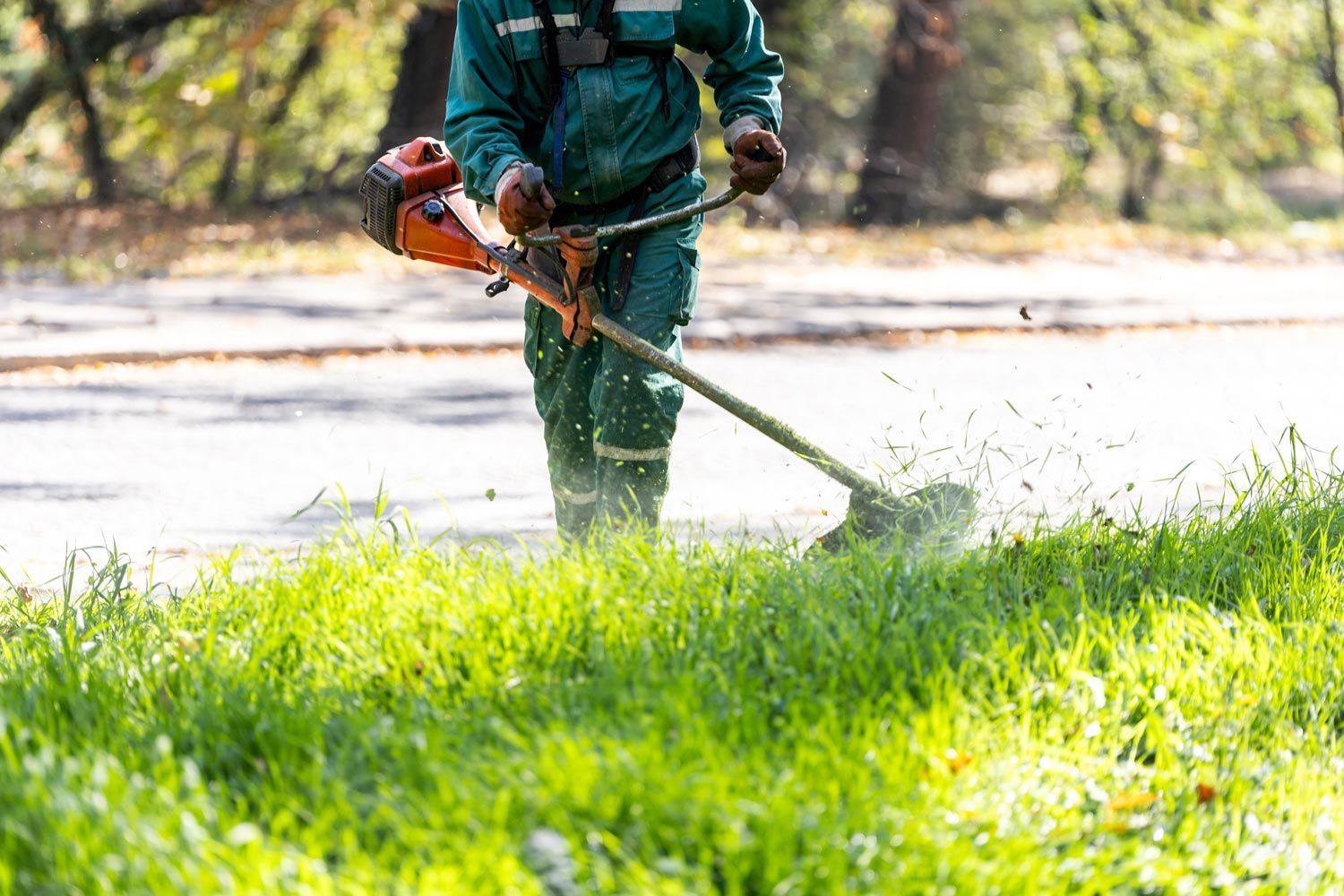 Person using a string trimmer to cut grass beside a path in a sunny park