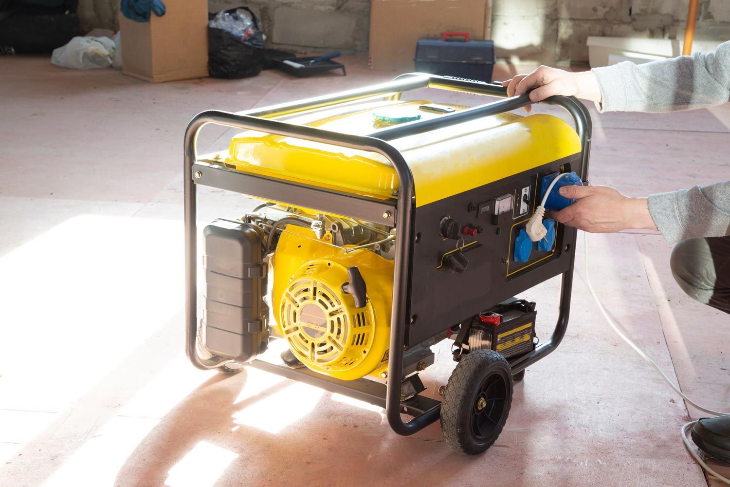 Yellow portable generator on a wheeled frame, with a hand turning its blue control knob indoors.