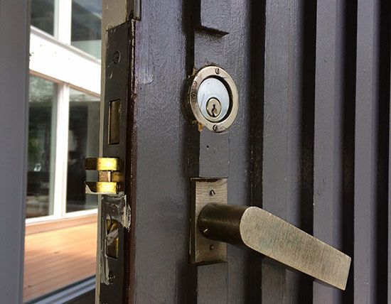 Close-up of a door lock and handle on a dark metal door near a glass wall.