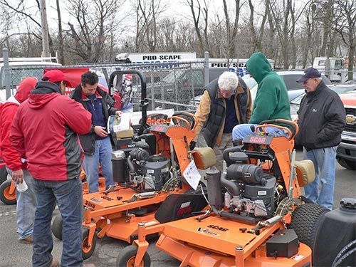 People gathered around orange lawn mowers at an outdoor equipment sale