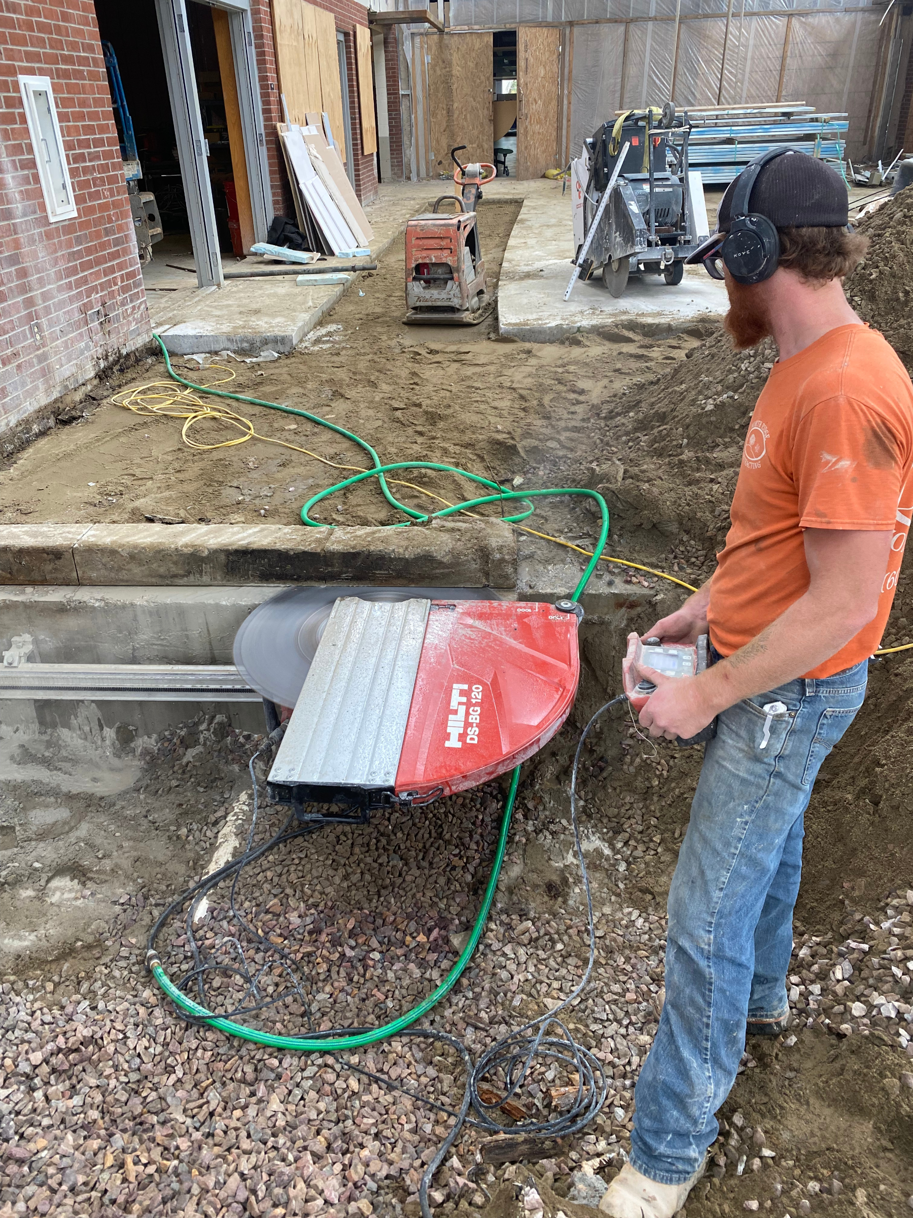 Construction worker using a concrete saw outdoors; red saw, green hose. He wears earmuffs and jeans.