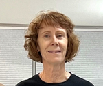 Smiling person with short curly hair wearing a black shirt, standing indoors against a light wall.