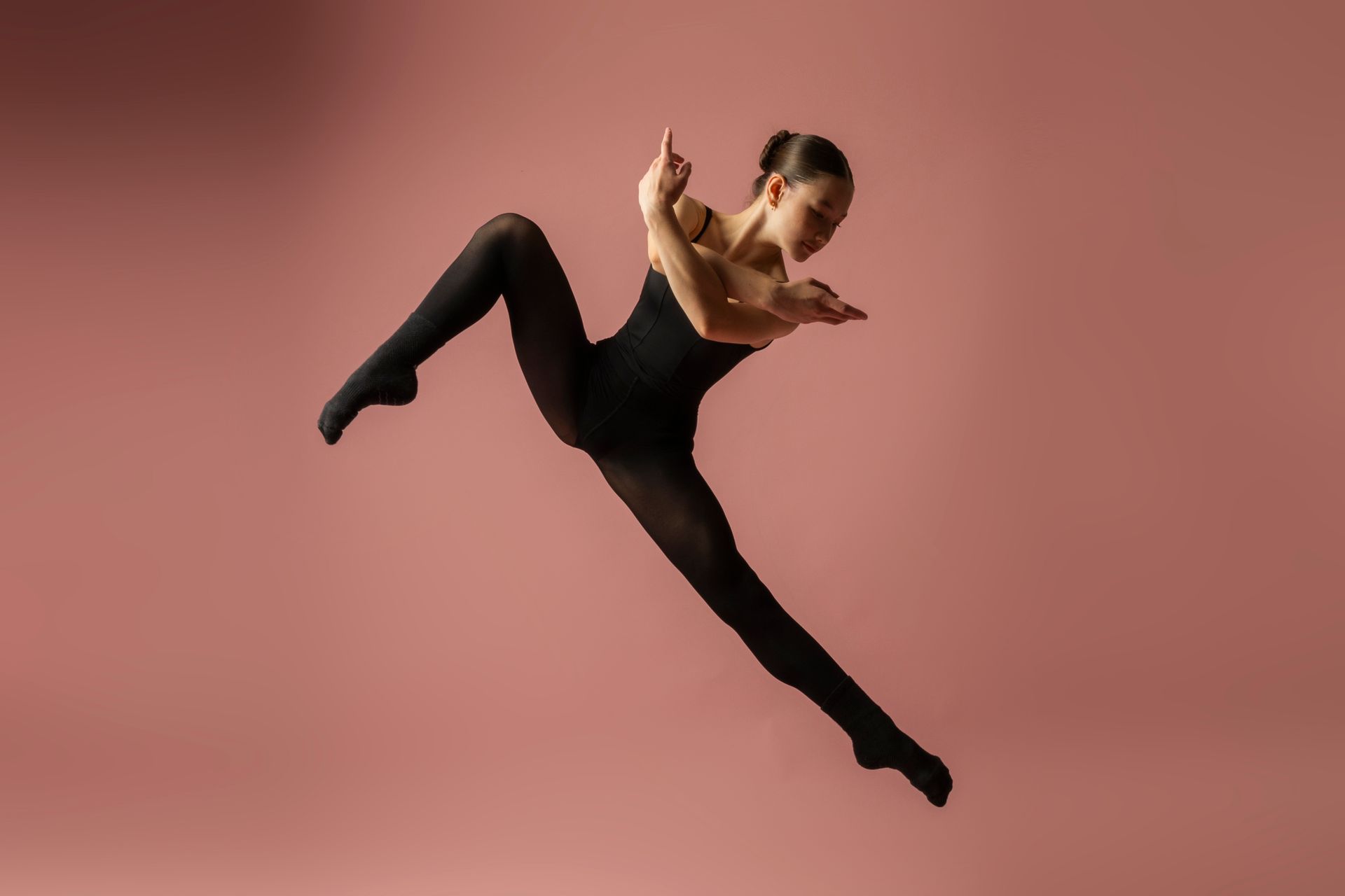 Dancer in black performs a high leap against a pink studio background