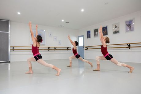 Three dancers in pink practice ballet lunges in a bright studio, arms raised overhead.