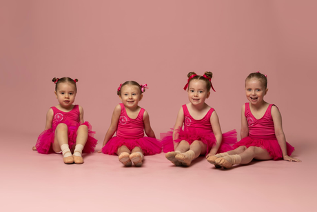 Four toddlers in matching pink outfits sit on a pink floor against a pink backdrop.