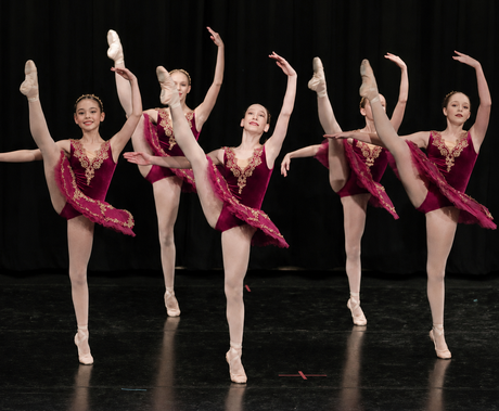 Five dancers in pink costumes perform high kicks on a stage.
