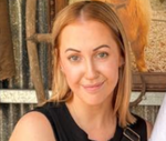Smiling person with blonde hair in a black top, standing outdoors near a rustic wall.