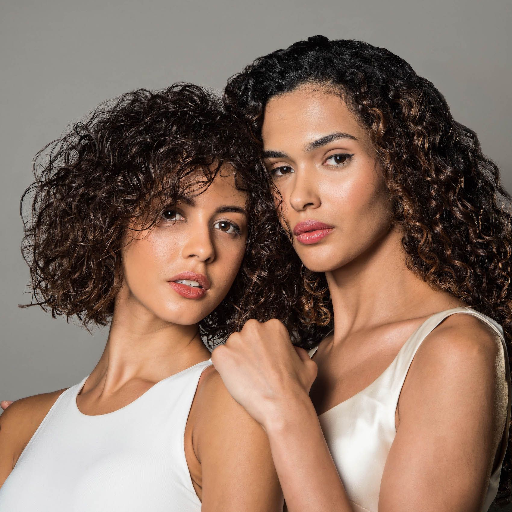 Due modelle con indosso top bianchi posano davanti a uno sfondo grigio, una con i capelli corti e ricci e l'altra con i capelli lunghi e ricci.