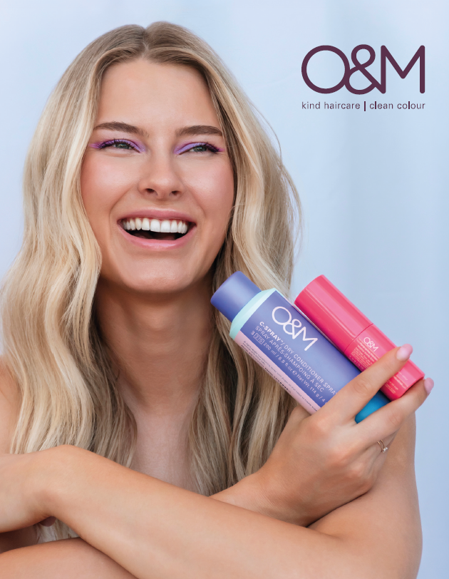 Una donna bionda sorridente tiene in mano due flaconi colorati di prodotti per capelli O&M su uno sfondo azzurro chiaro.