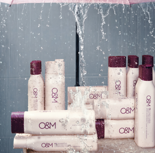 Flaconi e scatole per prodotti per la cura dei capelli O&M disposti su una superficie grigia e bagnata, con l'acqua che schizza alle loro spalle.