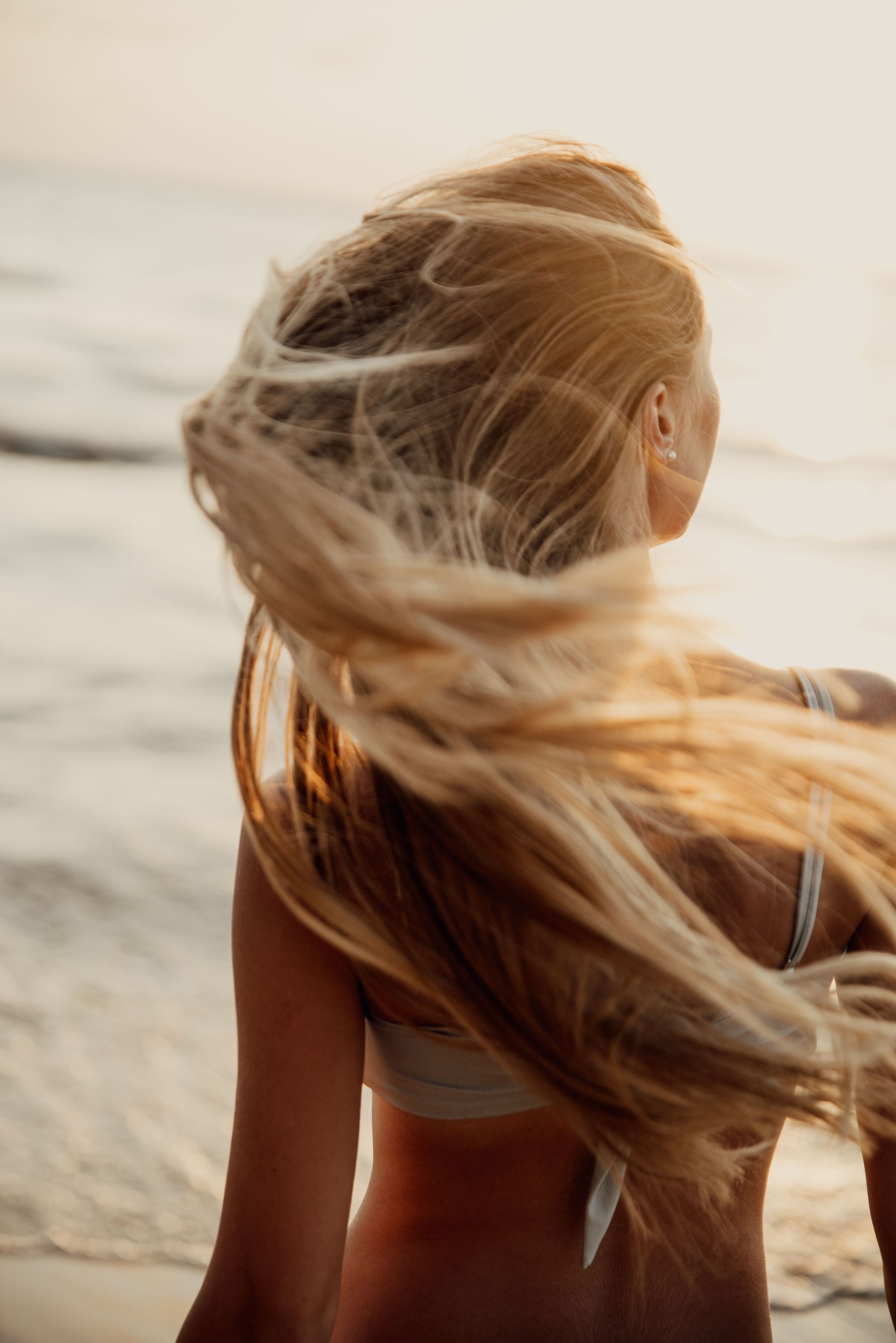 Persona dai capelli lunghi in piedi su una spiaggia, rivolta verso l'oceano, con i capelli mossi dal vento.