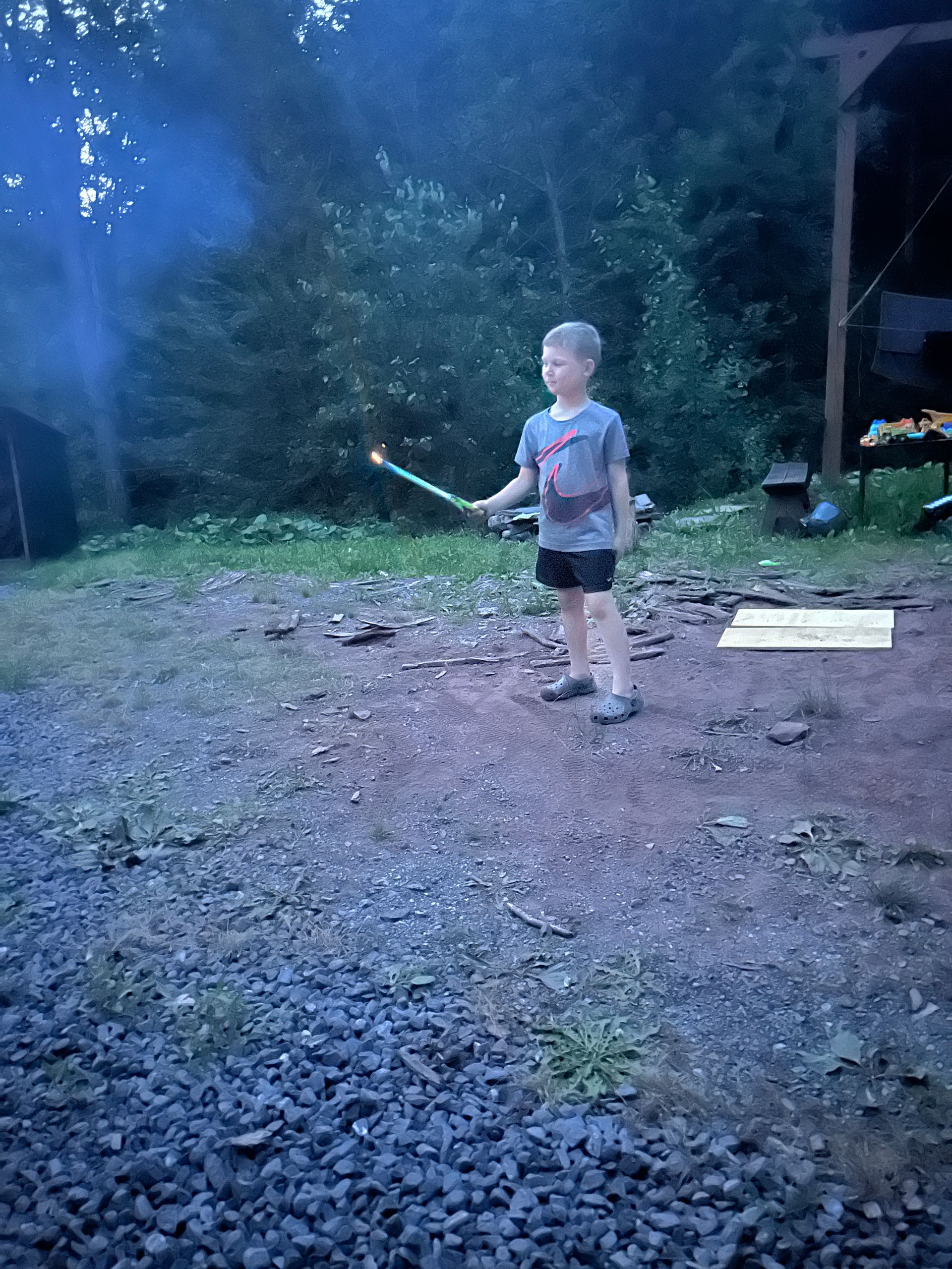 Child holding a glowing wand in a dim blue outdoor yard at dusk, standing on gravel near a porch