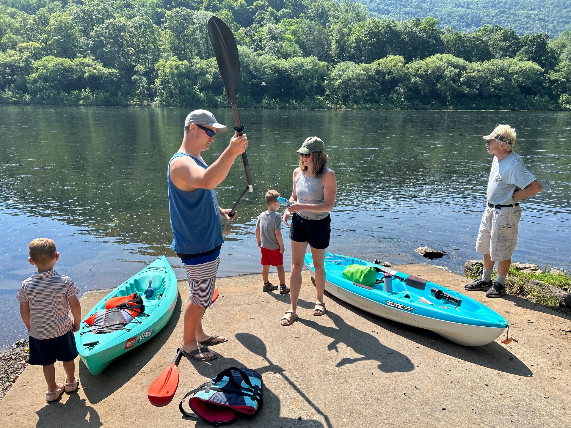 People by kayaks at a lakeshore, one holding a paddle, with a forested hill in the background