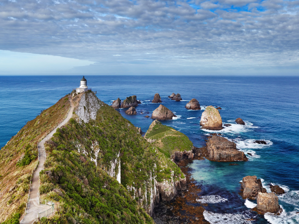Coastal cliff trail with a white lighthouse, green slopes, and blue ocean dotted with rocky islets.