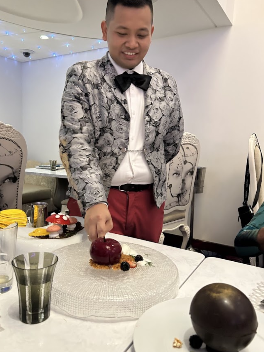 Person at a table arranging a dessert, with plated sweets and a large chocolate sphere nearby