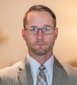 Man in suit and glasses looking directly at the camera with a neutral expression.