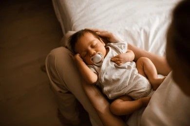 Bebé recién nacido durmiendo en el regazo, con pijama y chupete. Iluminación tenue, primer plano.