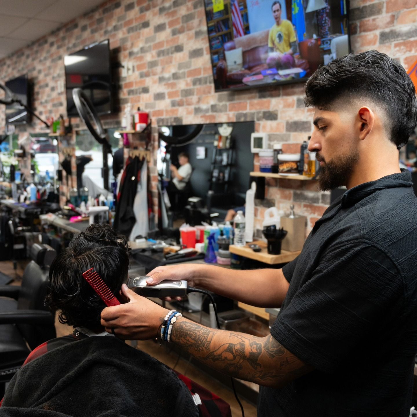 Barber trimming a customer’s hair in a busy brick-walled barbershop with mirrors and TVs.
