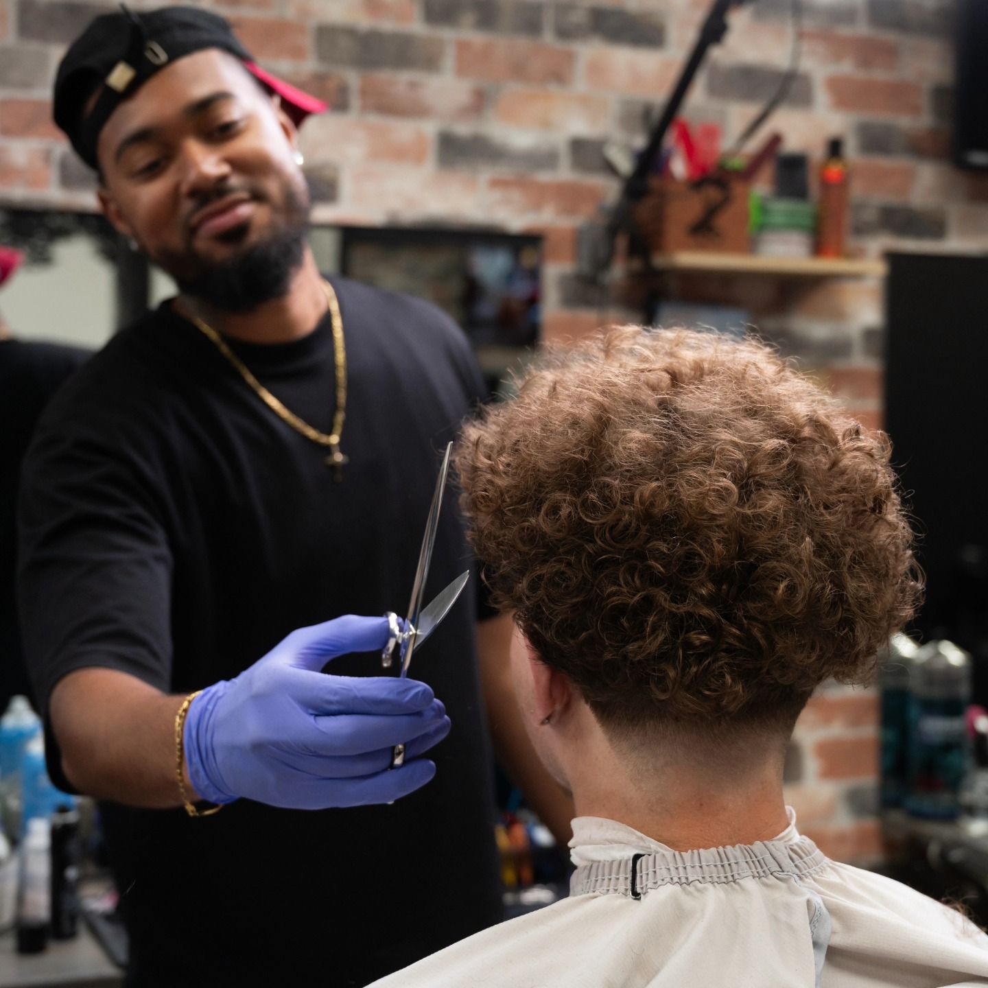 Barber in blue gloves cutting a client’s curly hair in a brick-walled barbershop