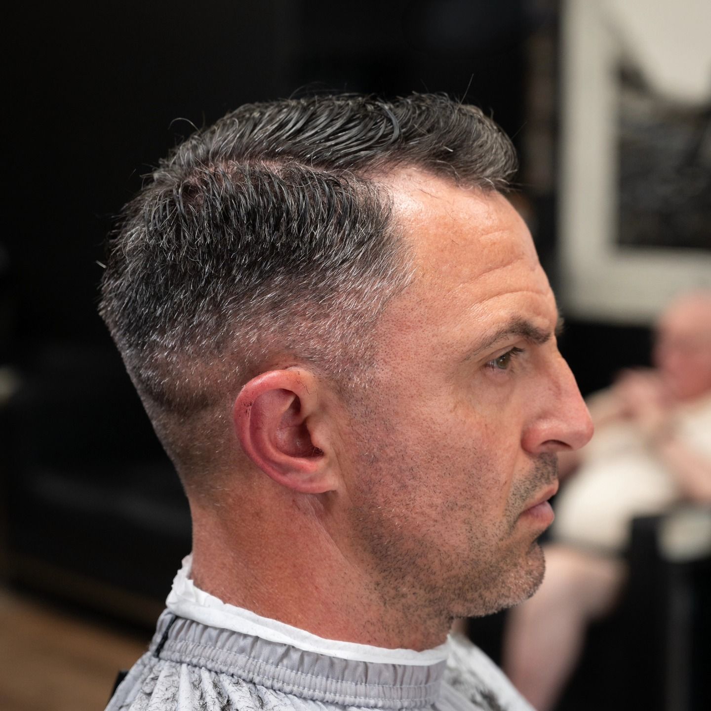 Side-profile of a man with a short fade haircut in a barber chair, wearing a striped cape.