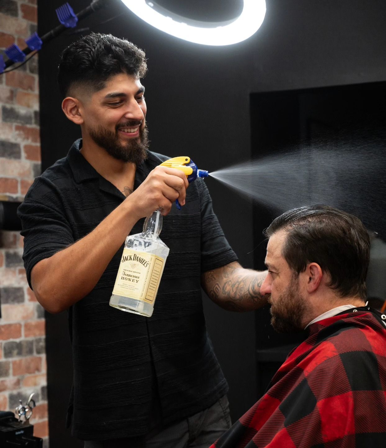 Barber spraying hairspray on a seated client in a barbershop, smiling and chatting.