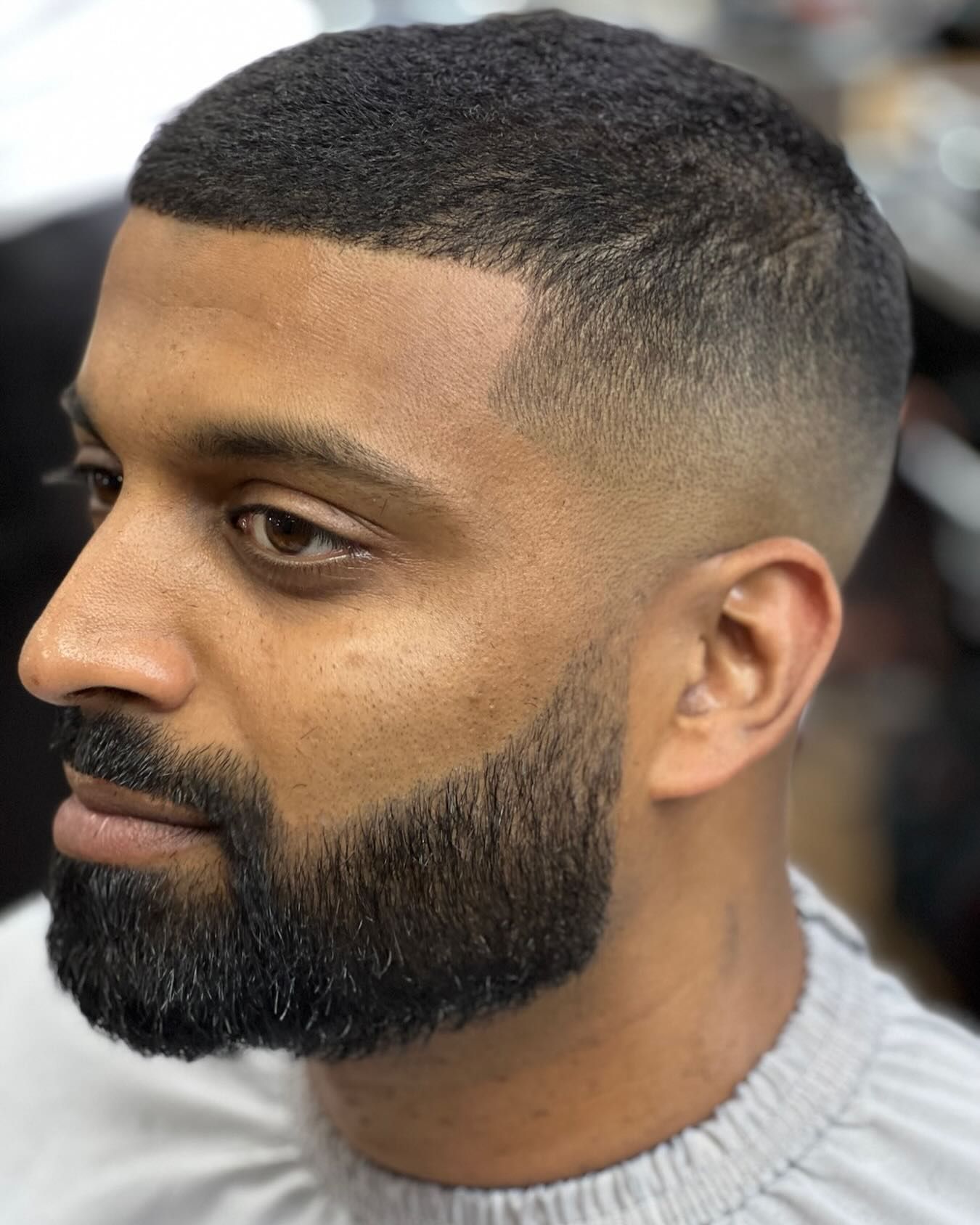 Side-profile of a man with a short fade haircut and full beard in a barbershop chair.
