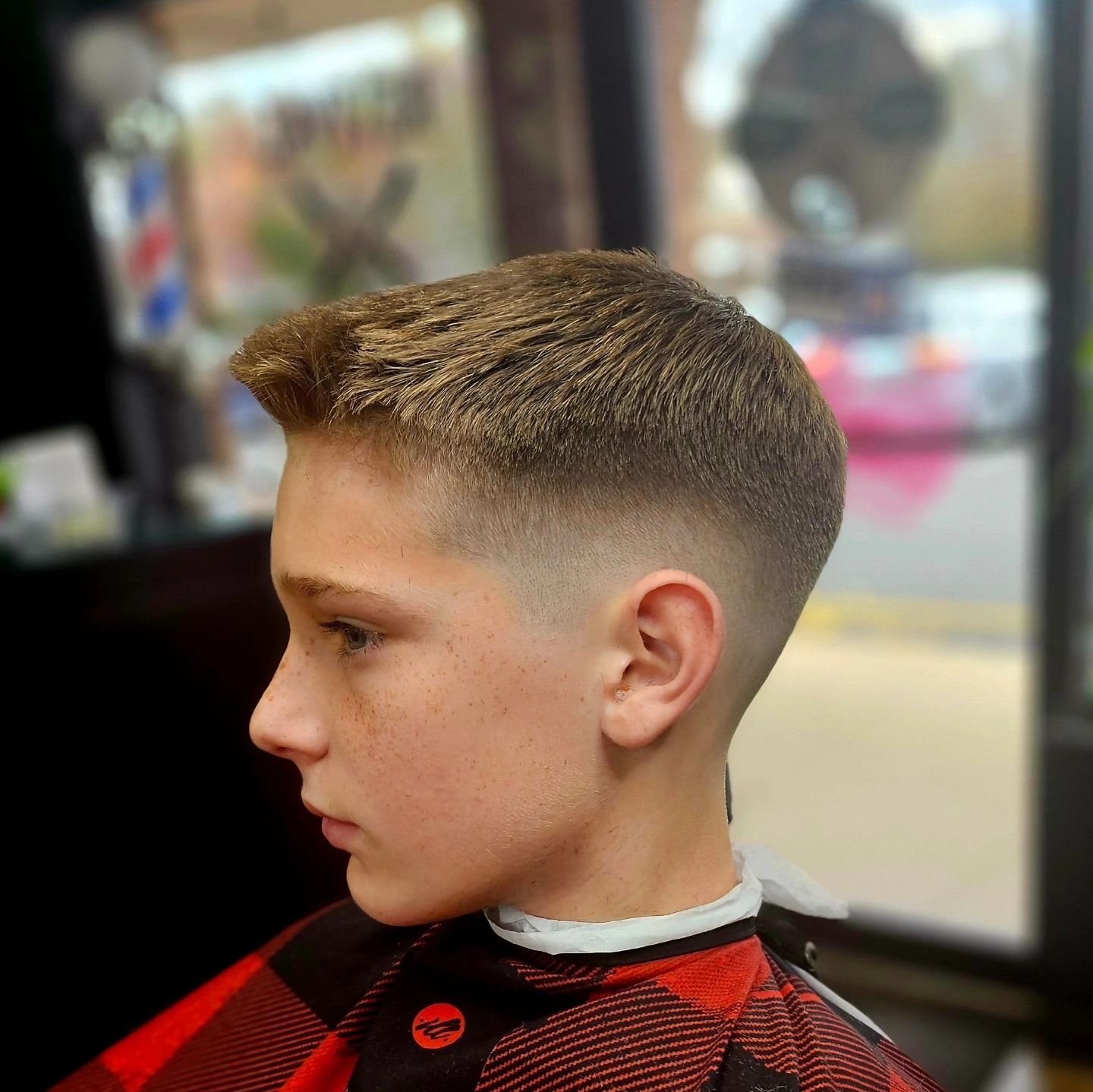 Boy with a short fade haircut in a red-and-black plaid shirt, sitting in a barbershop chair.
