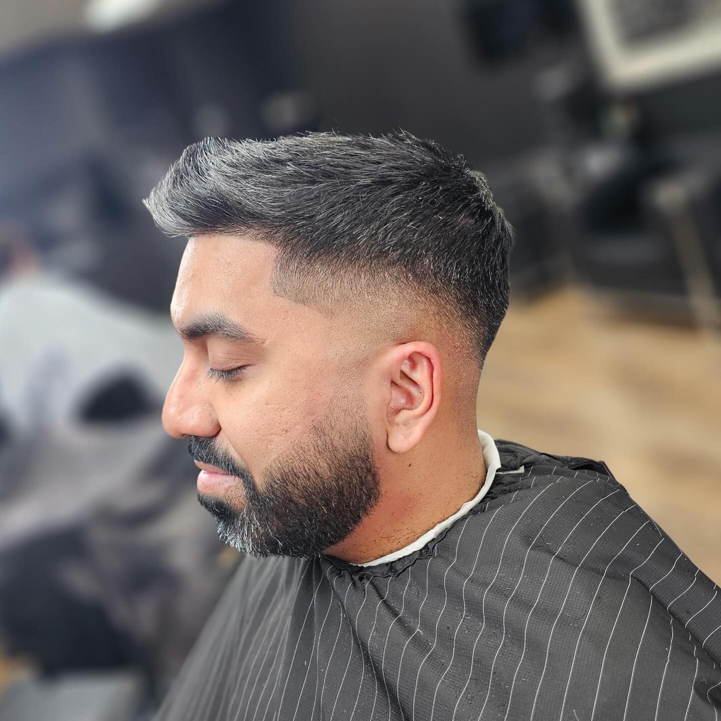 Side profile of a man with a fade haircut and beard in a barber chair, wearing a black striped cape.