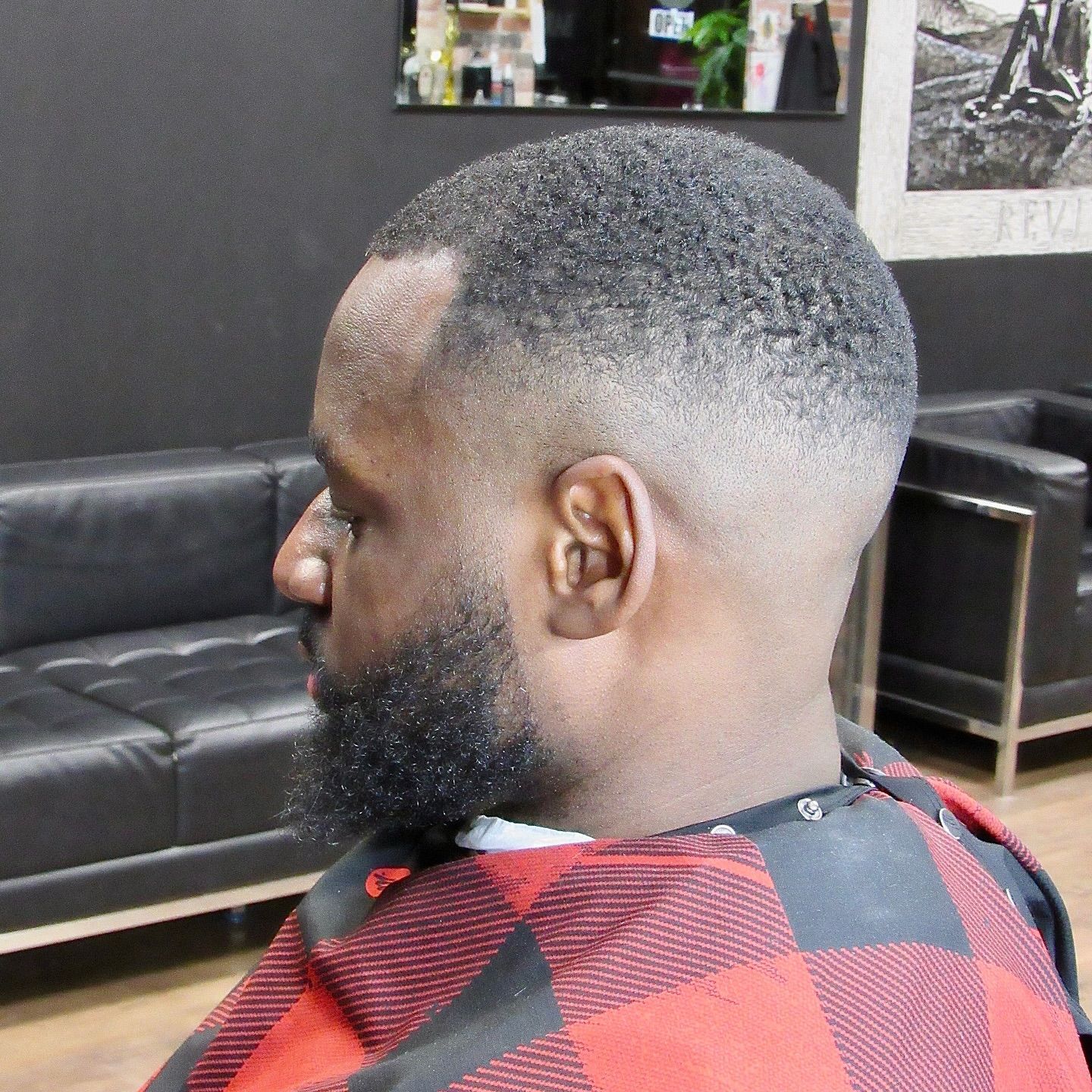 Man getting a fresh fade haircut in a barbershop, seated in a black chair with red plaid cape.