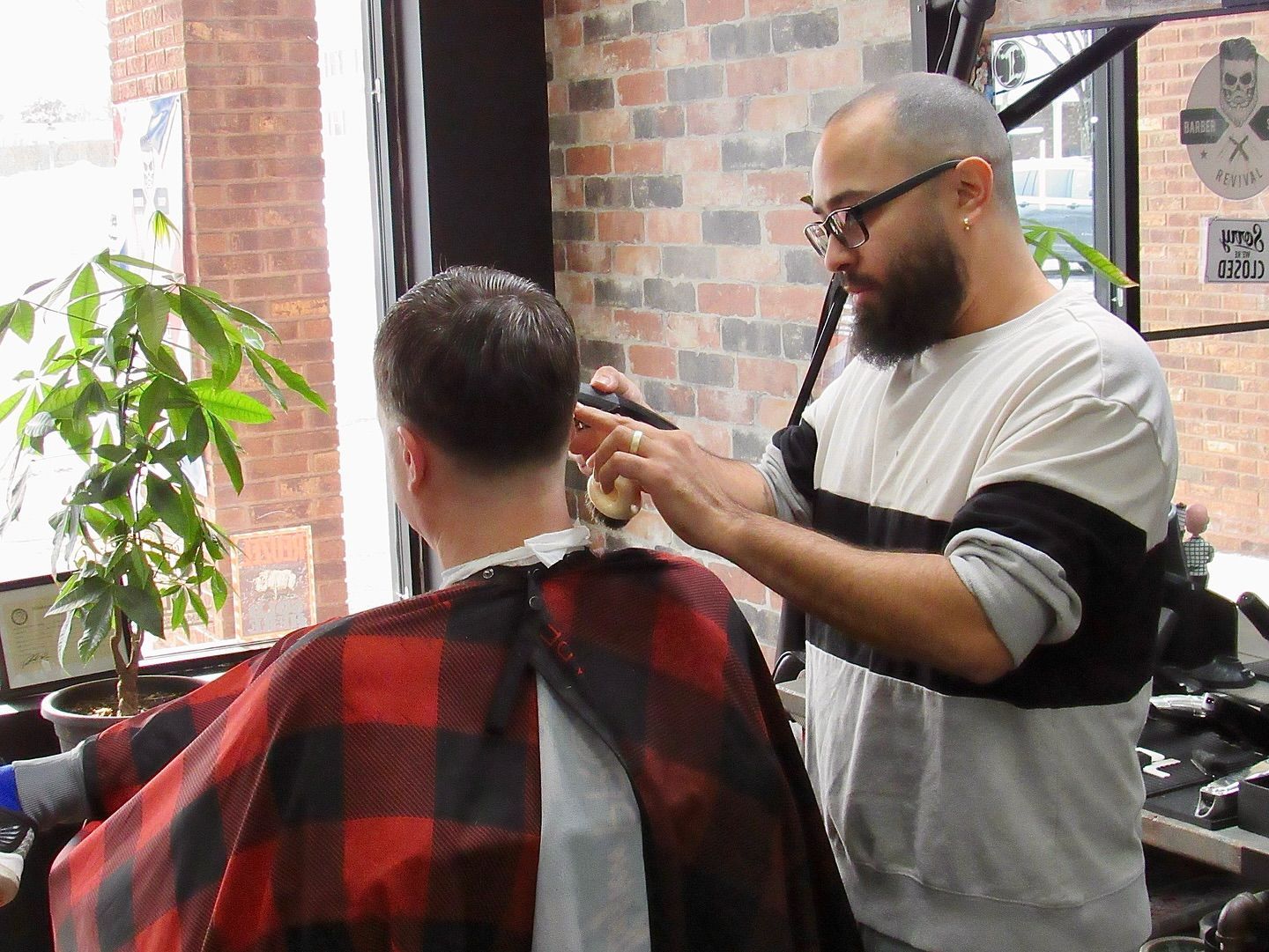 Barber cutting a customer’s hair in a barbershop, with a red-and-black cape and brick wall background