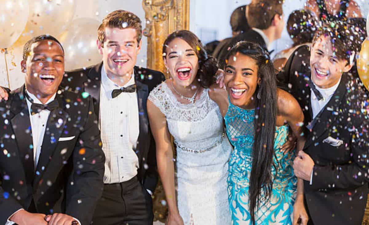 A group of smiling teens at a prom, confetti falling. People are wearing formal wear, standing in a decorated hall.