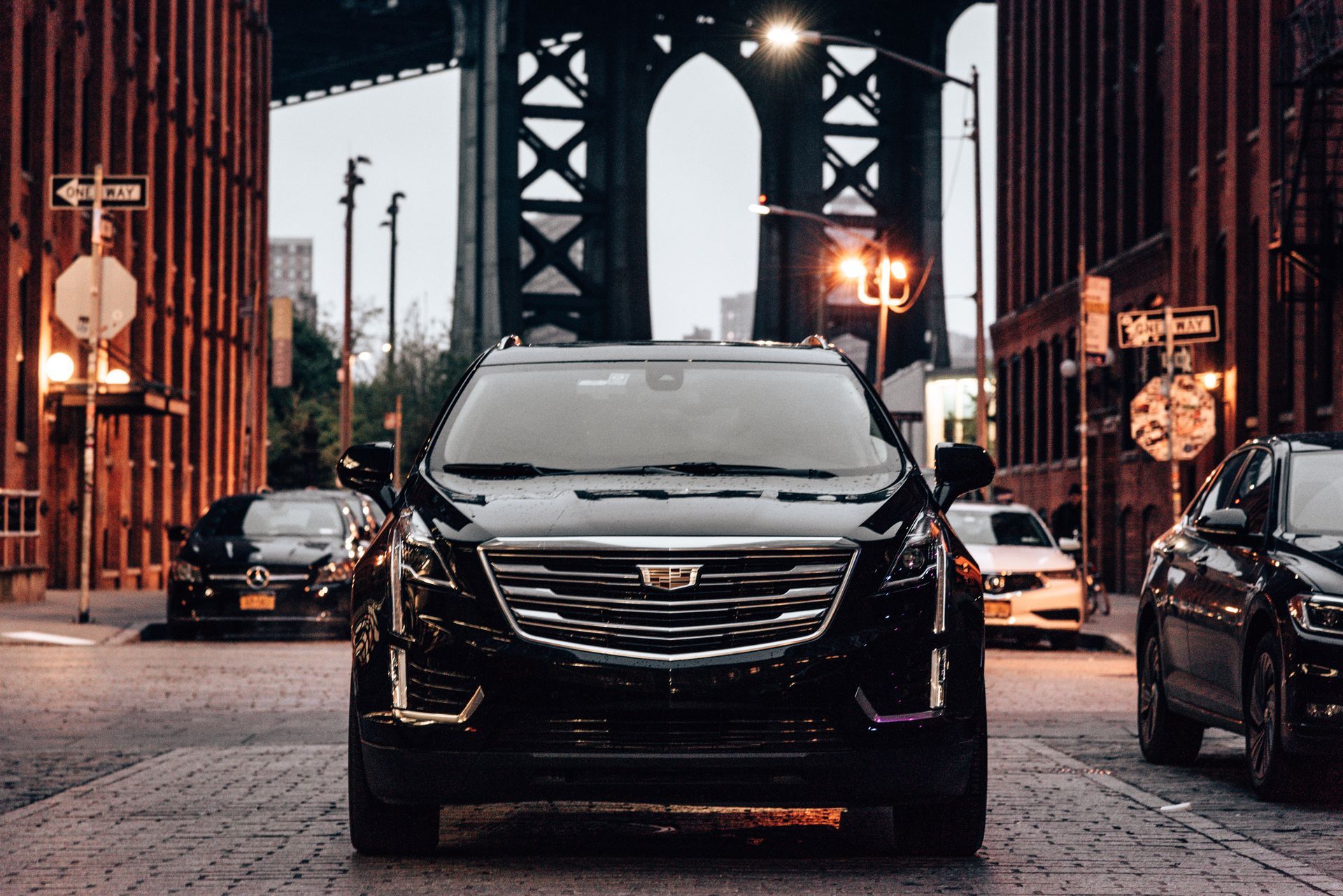 A black Cadillac SUV parked on a cobblestone street in front of the Manhattan Bridge. Other cars and buildings surround it.
