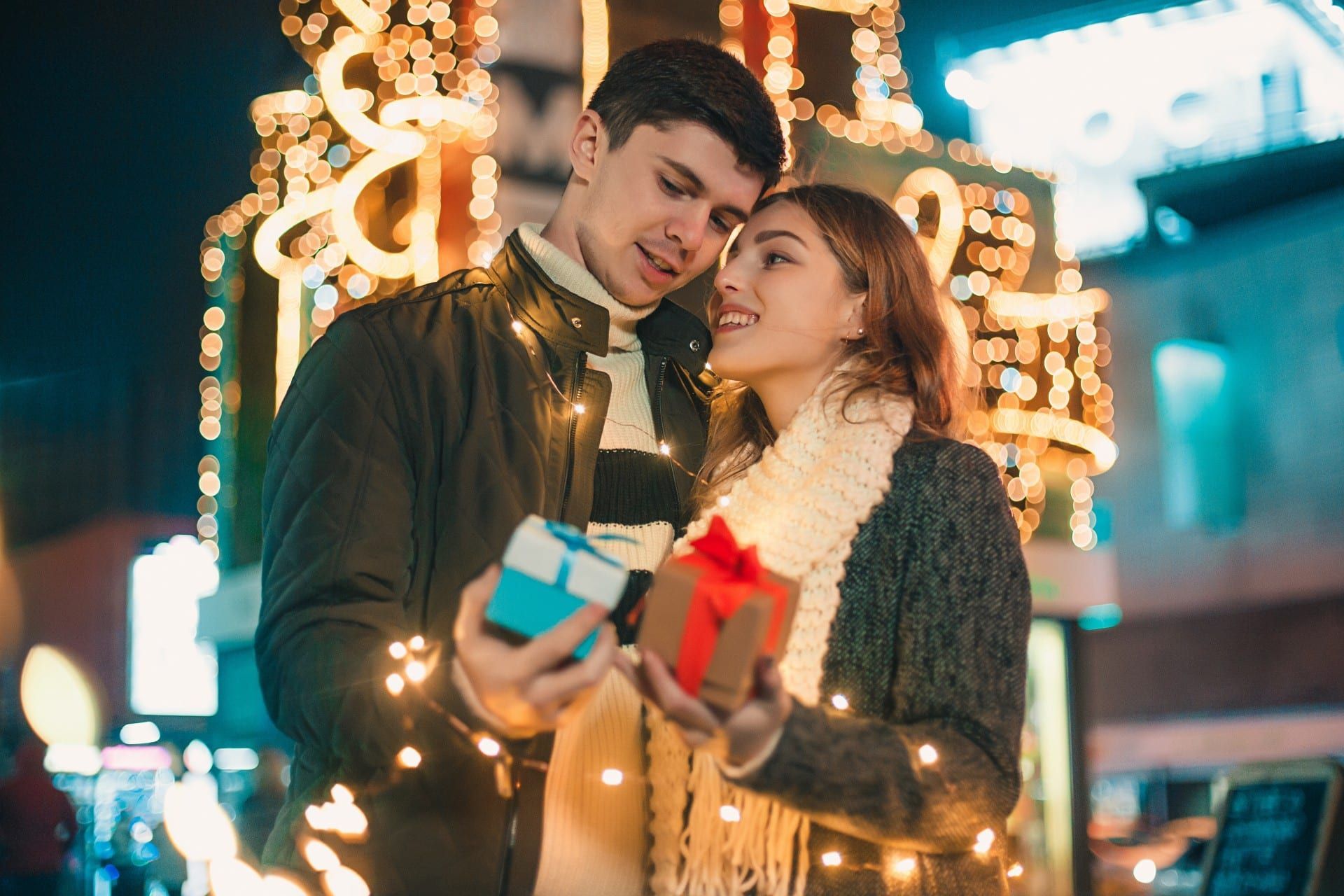 A couple exchanging gifts outdoors at night, surrounded by festive lights.