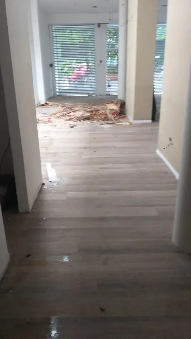 Pasillo vacío con suelo de madera clara y una ventana que deja ver escombros en el suelo cerca del fondo.