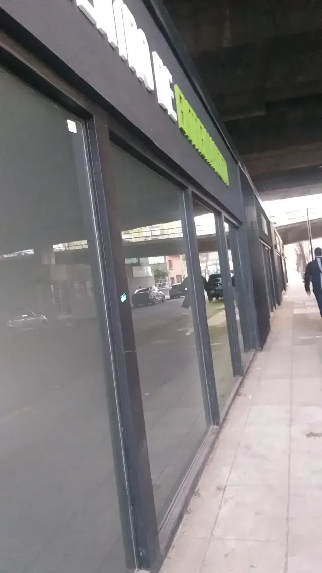Acera cubierta frente a las tiendas con ventanas de vidrio oscuro y un toldo negro con letreros verdes.