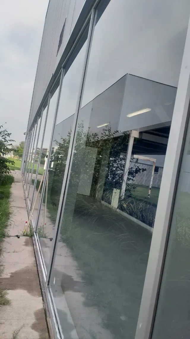 Entrada a un edificio moderno con fachada de cristal, ventanas reflectantes y una acera junto a zonas verdes.