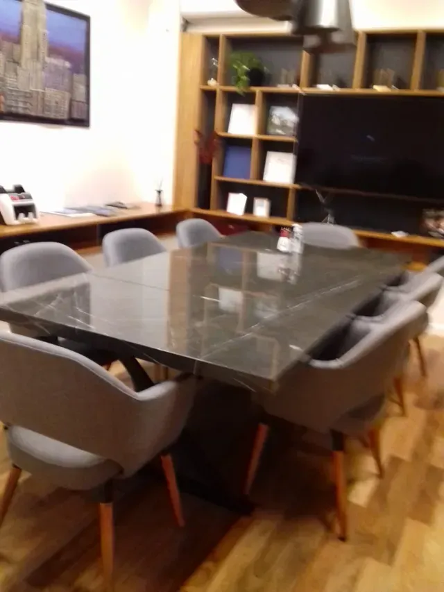Comedor con mesa de cristal, sillas grises y un televisor grande adosado a un mueble de pared.