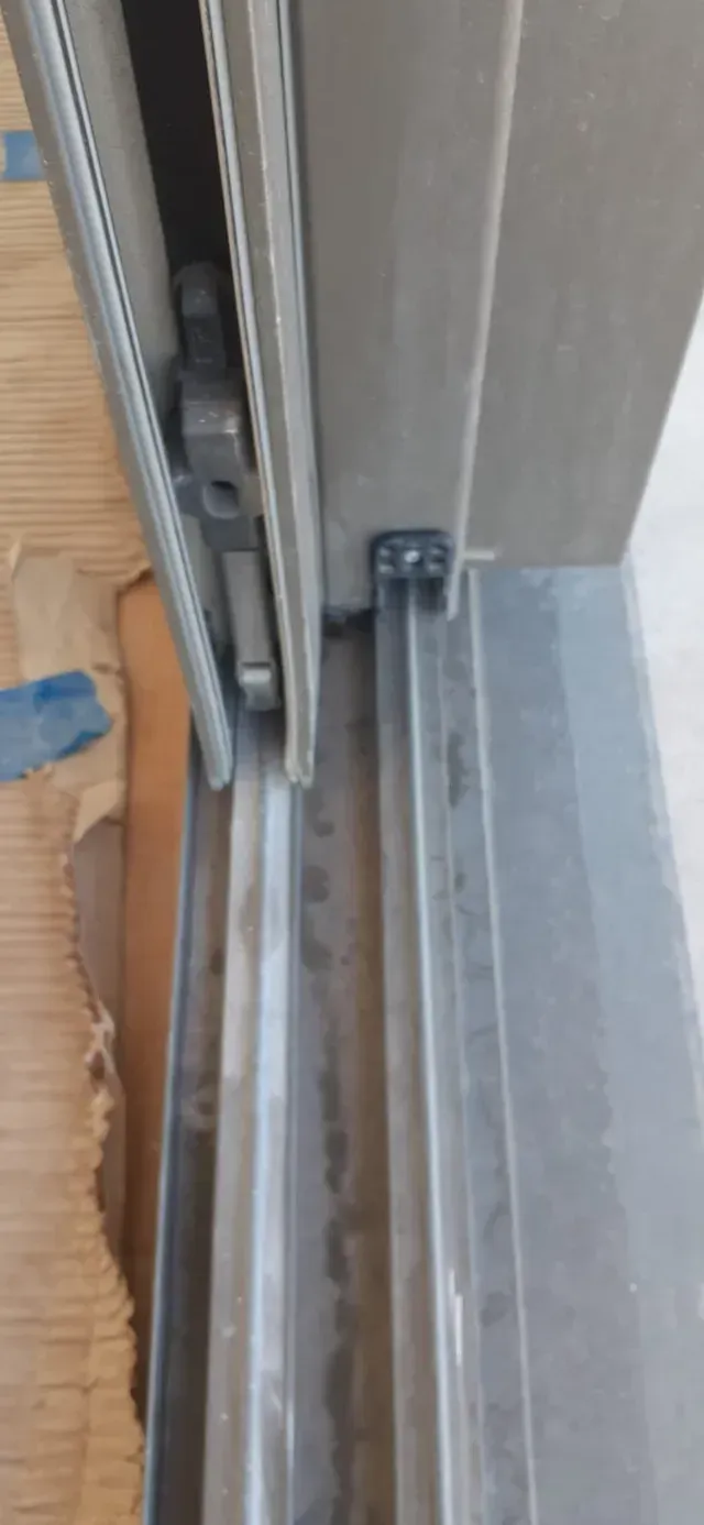 Primer plano de un riel y pestillo de puerta corrediza en un marco gris con piso de madera cerca.