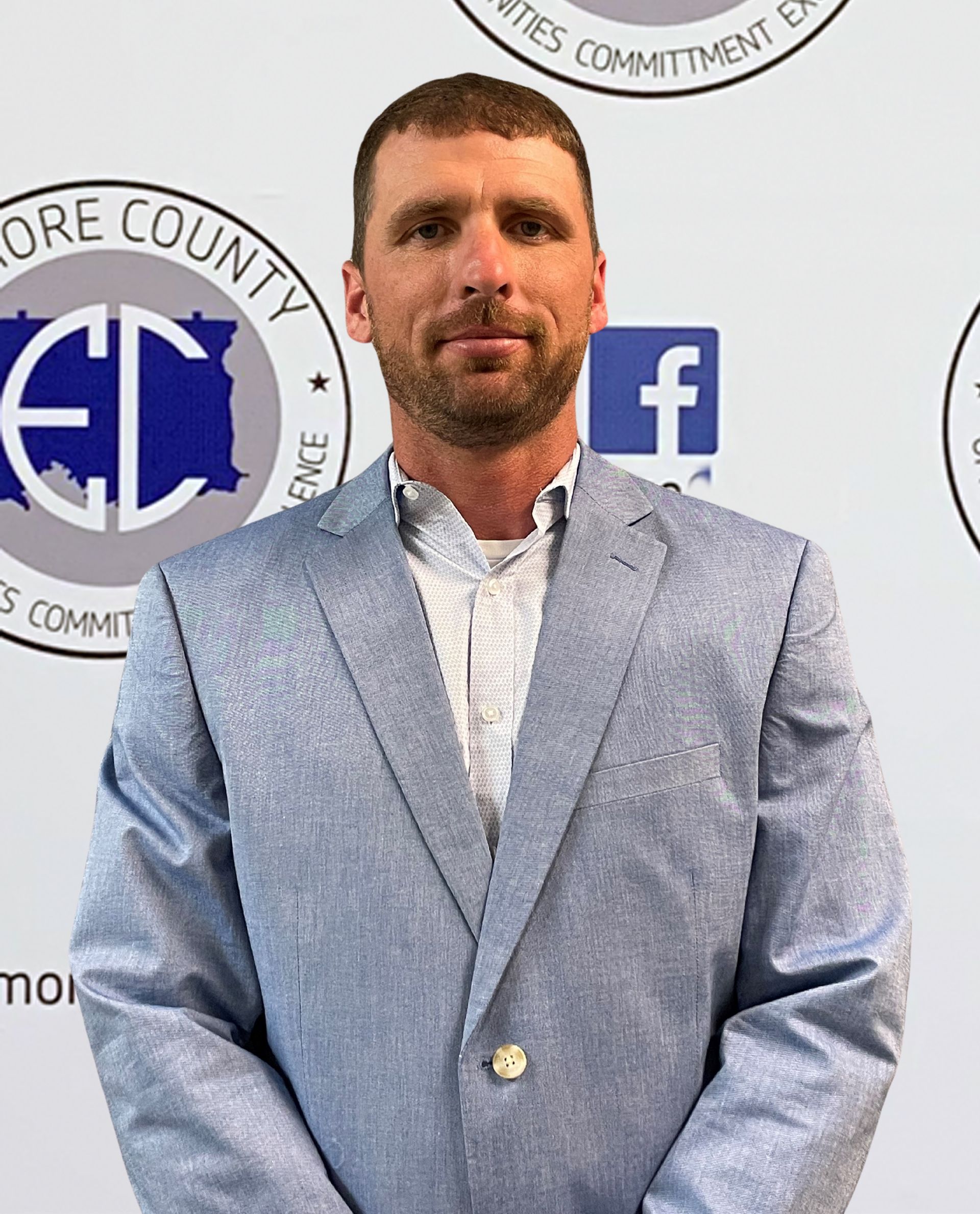 Man in a light blue blazer and white shirt stands in front of a backdrop with logos. He is looking directly at the camera.