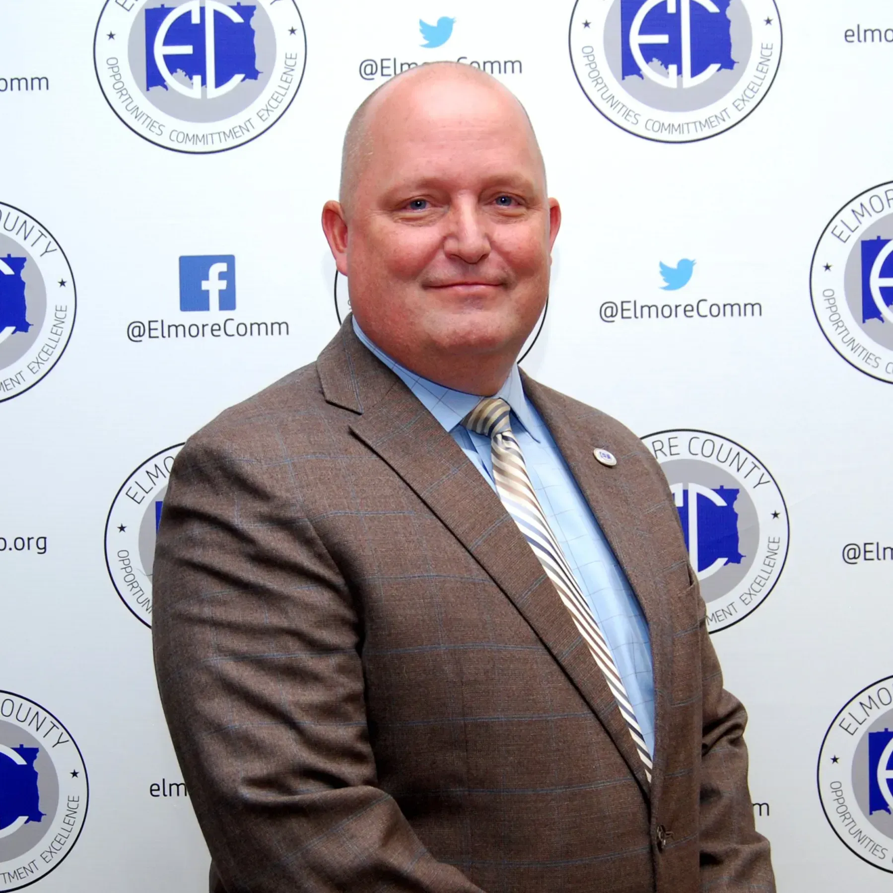 A bald man in a brown suit stands in front of a backdrop with a logo. He smiles and wears a tie.