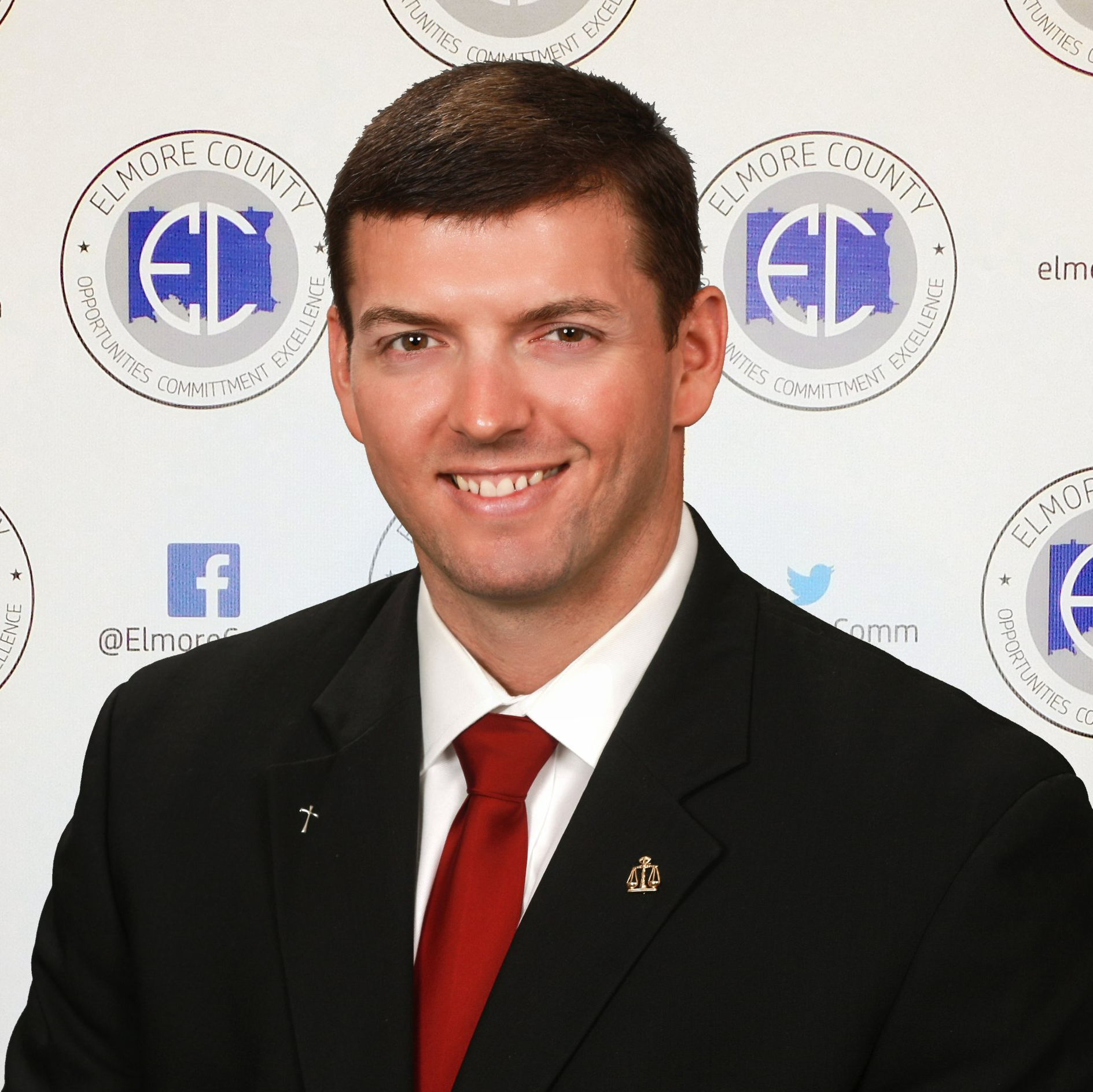 Man in a black suit and red tie smiling. He is in front of a backdrop with logos.