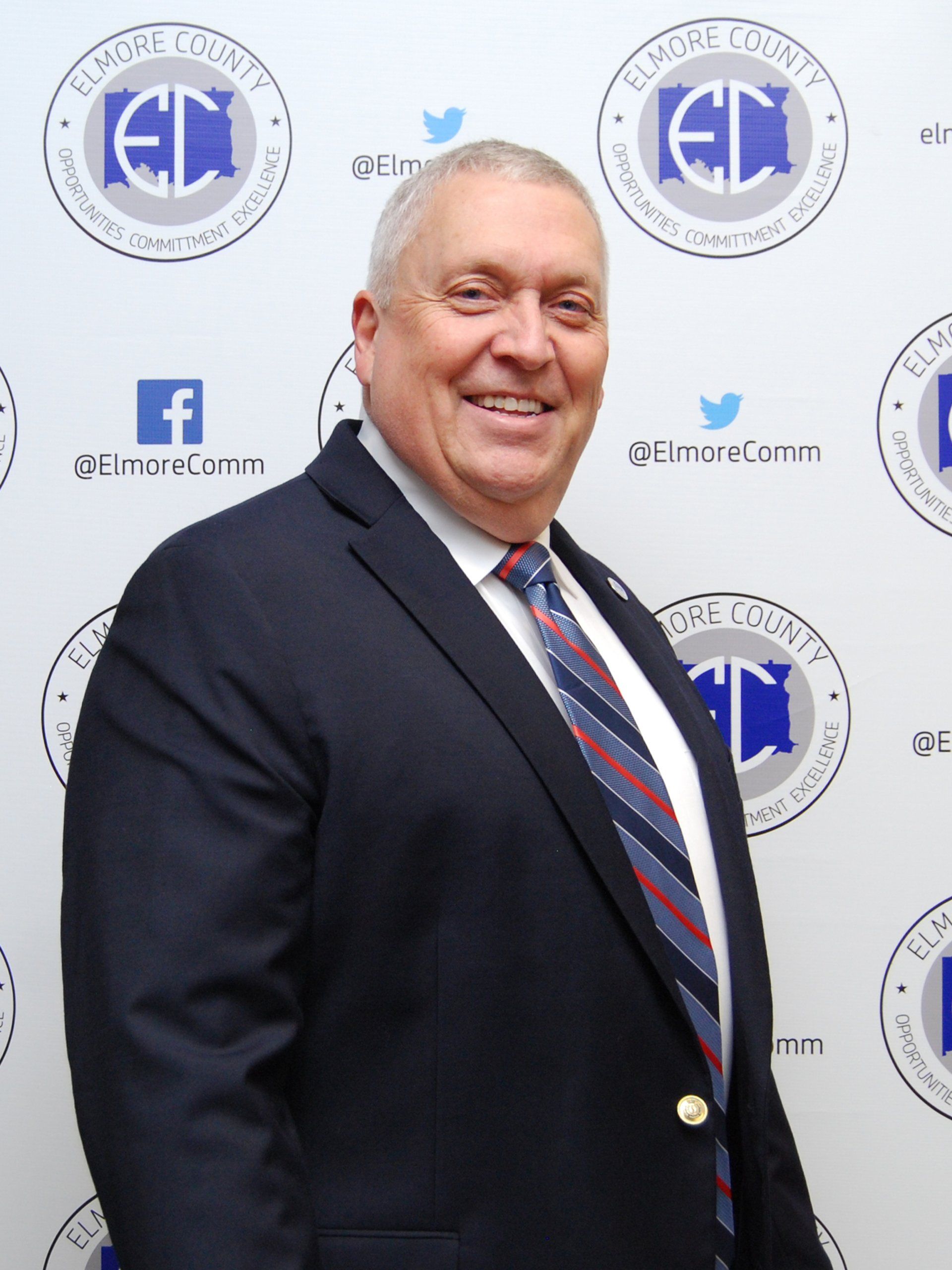 Man in a dark suit and tie, smiling at the camera in front of a logo-patterned background.