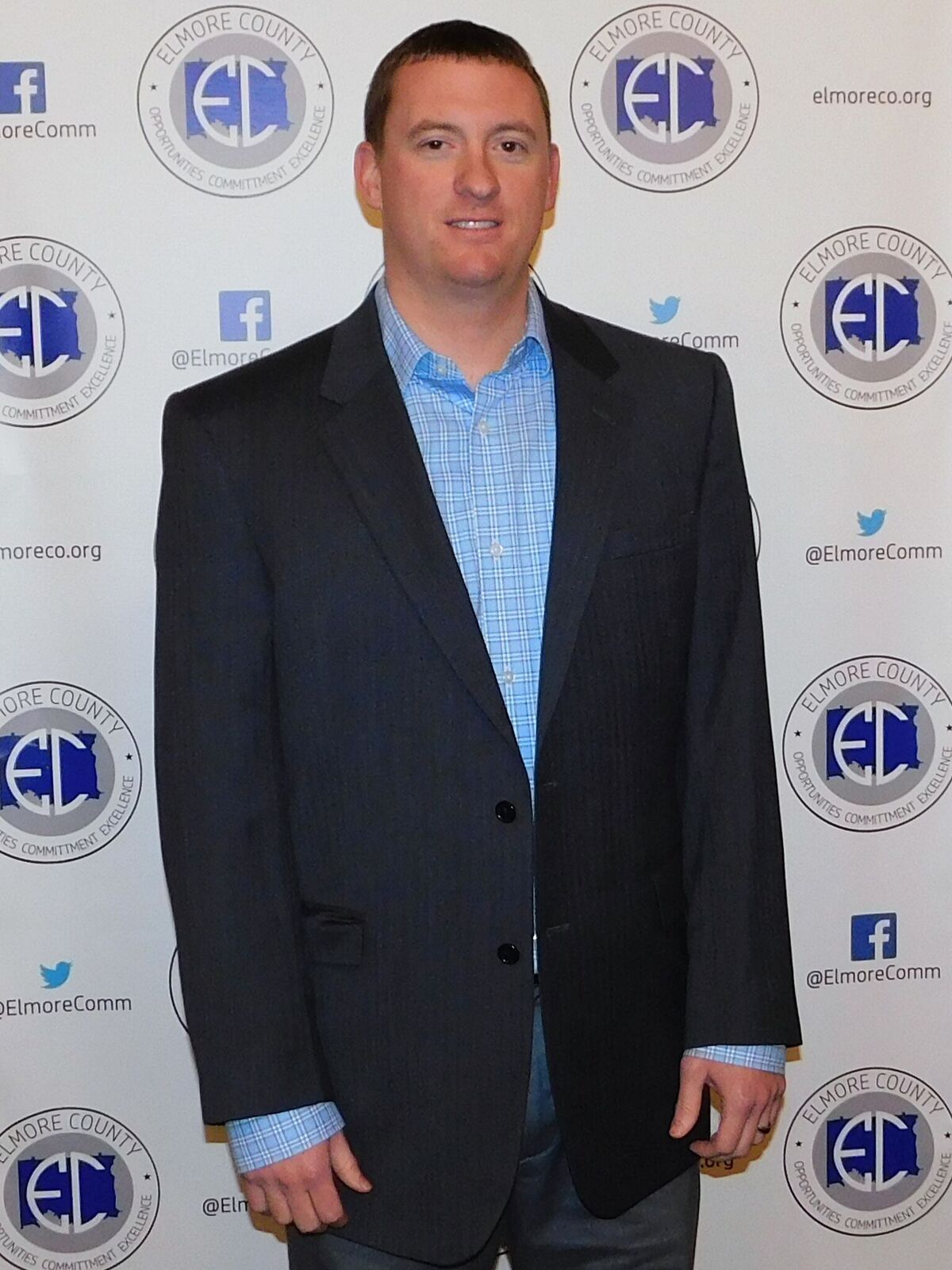 A man in a dark suit and blue patterned shirt stands in front of a backdrop with logos.