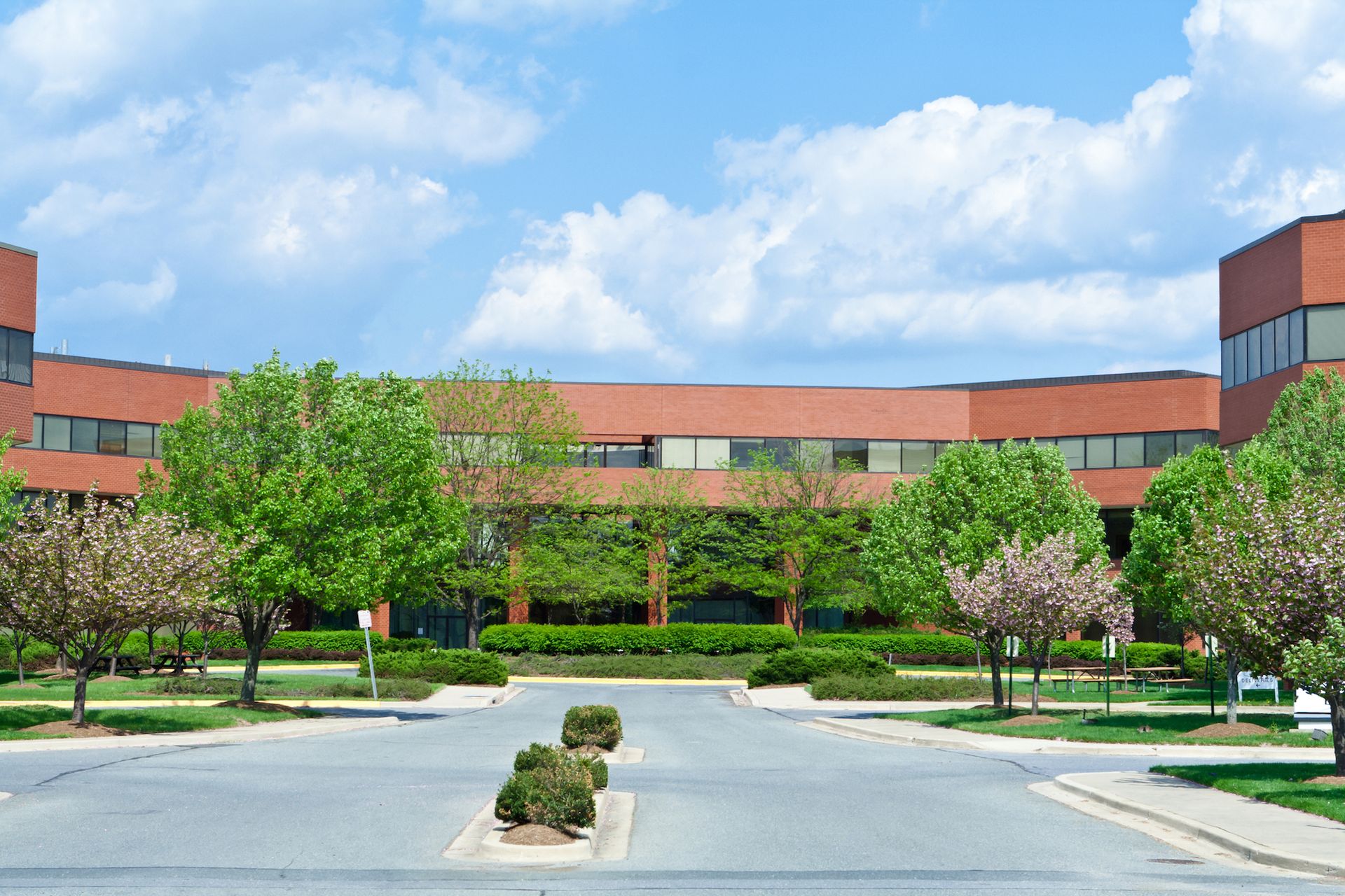 Brick office building with a curved facade, green trees, blue sky, and a paved entrance.