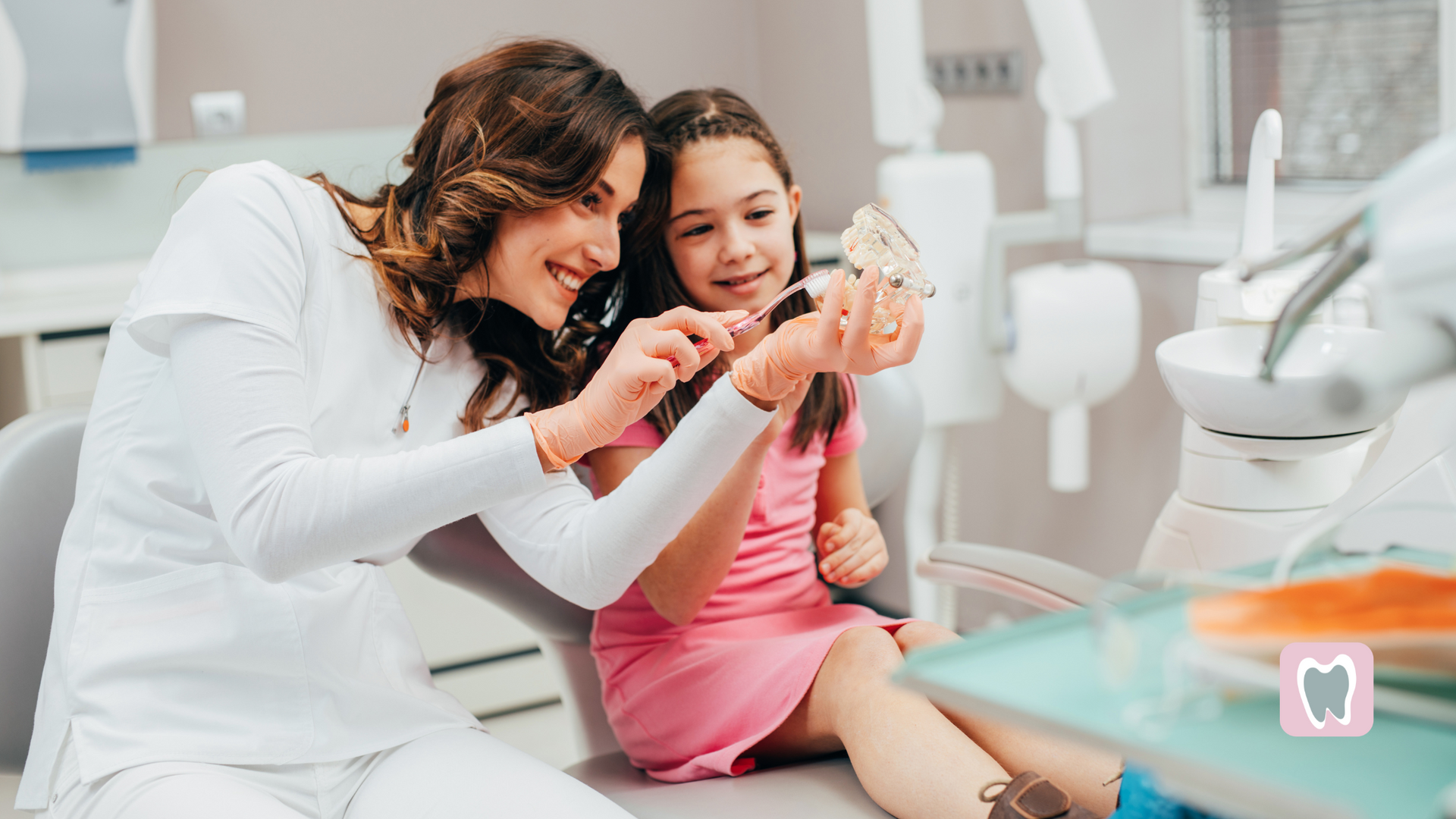 A woman and a little girl are sitting in a dental chair looking at a tooth model.
