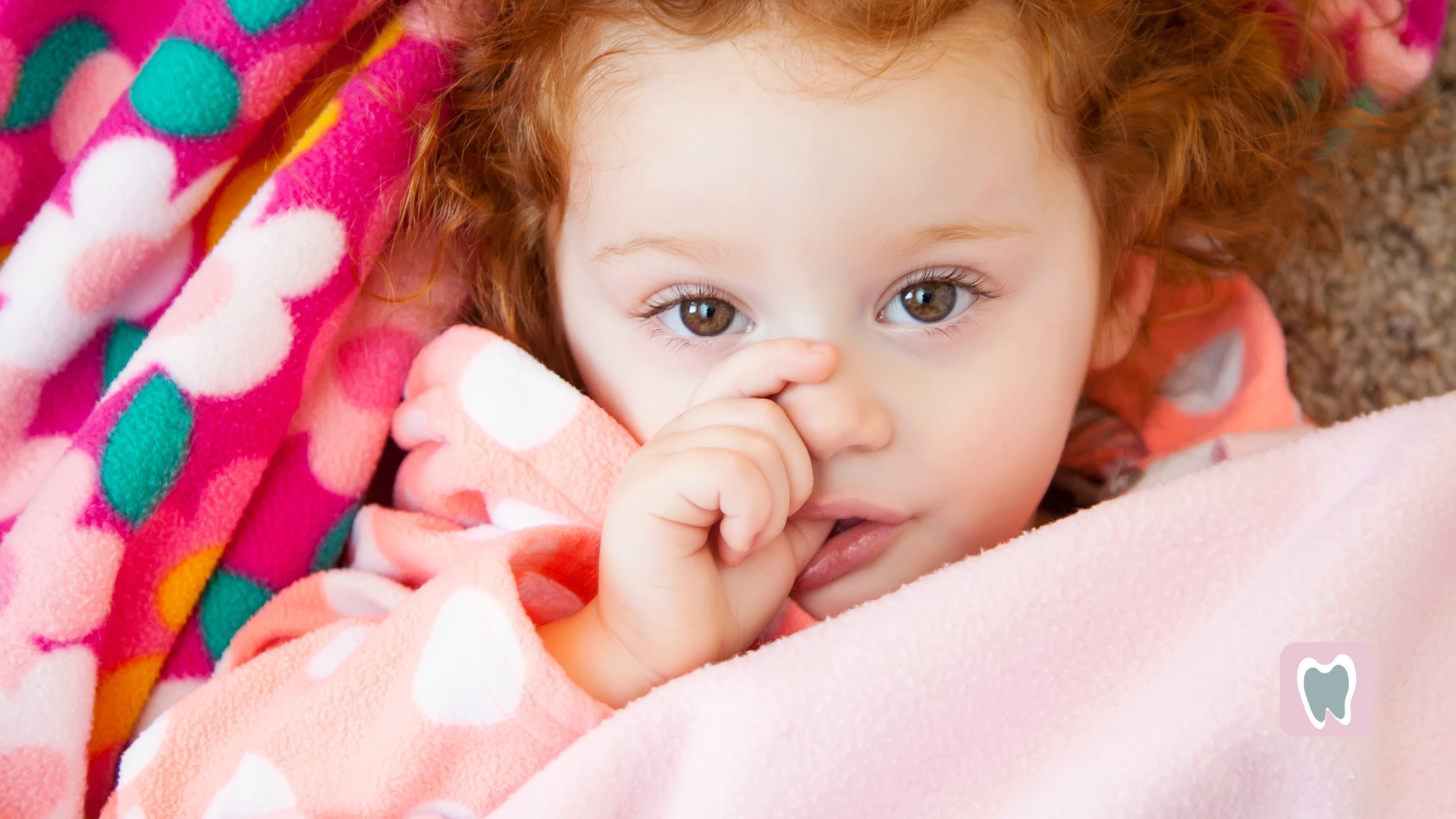 A little girl is laying under a blanket with her finger in her nose.