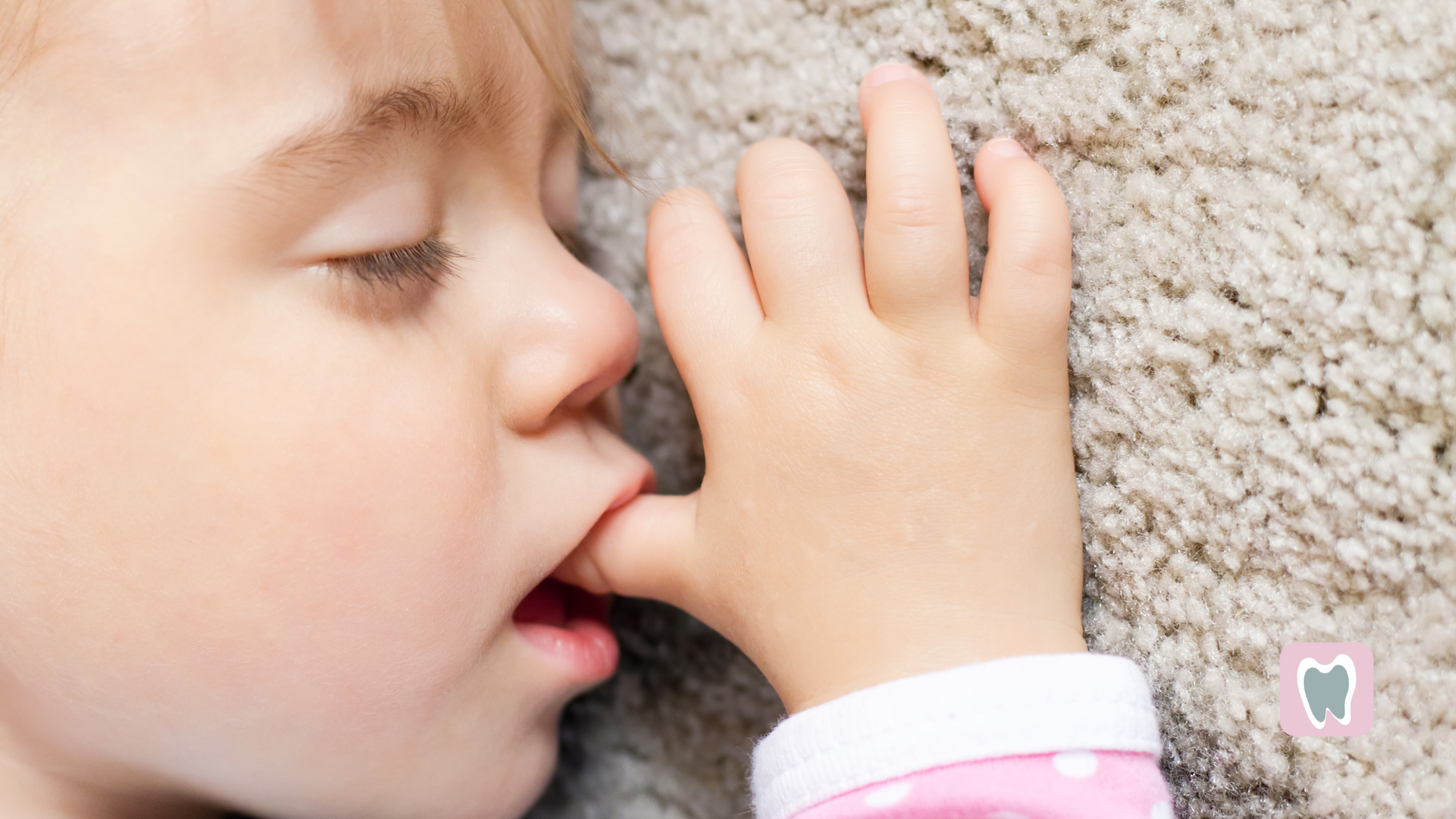 A little girl is sleeping on a carpet and biting her thumb.